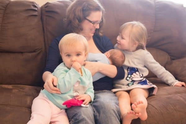 Mother breastfeeding newborn baby on sofa with preschooler and toddler daughters sitting with her