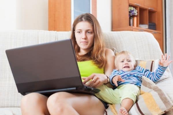 Mom sitting beside crying toddler, trying to work on laptop and feeling frustrated