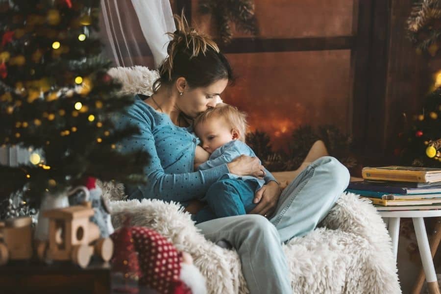 Toddler sitting in Mommy's lap breastfeeding beside Christmas tree and fireplace