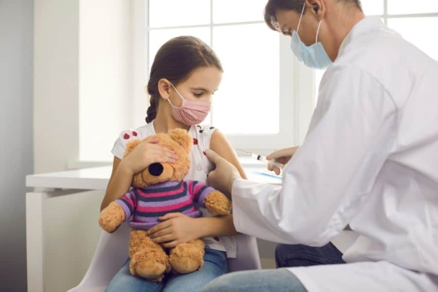 little girl holding a teddy bear and wearing a mask before she gets a shot