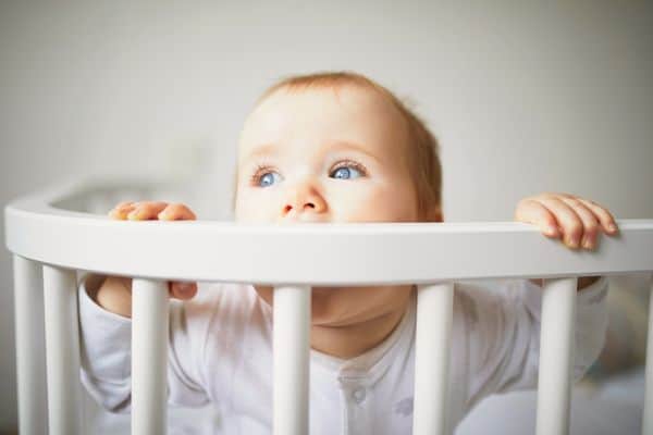 Baby girl chewing on her crib railing while standing at the side of her crib.