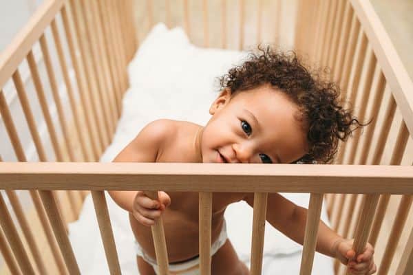 Little boy standing in his crib. The crib mattress is lowered to the bottom and there are no pillows or toys in his crib to climb on, so he is safe from falling out.