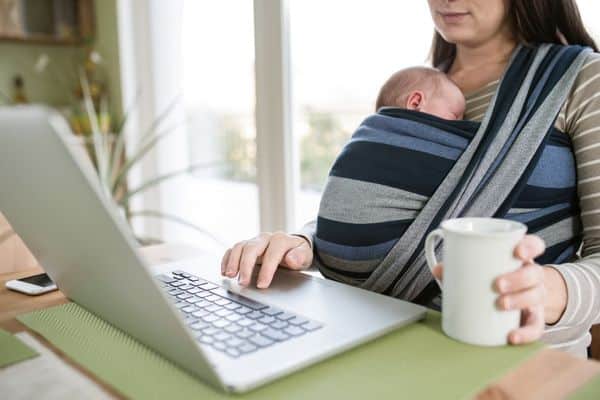 Woman working at laptop while baby is sleeping on her in soft carrier